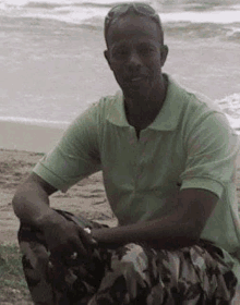 a man in a green polo shirt and camouflage pants is sitting on the beach .