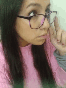 a woman wearing glasses and a pink sweater holds her finger to her nose