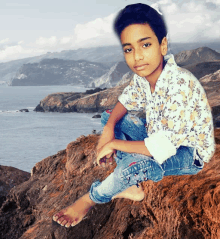 a young boy sits barefoot on a rock near a body of water