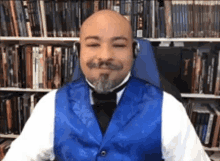 a bald man with a beard and mustache wearing a blue vest and bow tie is sitting in front of a bookshelf .