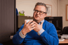 a man wearing glasses and a blue sweater sitting in front of a dell monitor