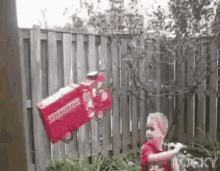 a little boy is standing in front of a wooden fence with a red fire truck hanging from it .