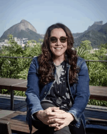 a woman wearing sunglasses and a denim jacket sits with her hands folded