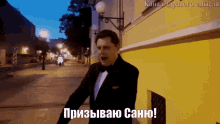 a man in a tuxedo is walking down a street at night and says " призываю саню "