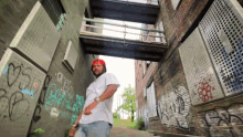 a man in a red hat stands in an alleyway with graffiti on the walls including one that says ' gray '