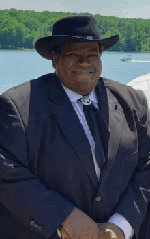 a man wearing a cowboy hat and a suit is standing in front of a body of water .