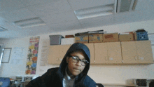 a boy wearing a hooded jacket and glasses in a classroom with a poster on the wall that says " how to win "