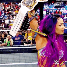 a woman with purple hair is holding a wrestling championship belt over her head in front of a crowd .