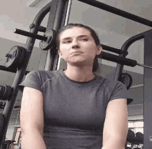 a woman in a gray shirt is sitting in front of a gym machine and looking at the camera .