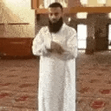 a man with a beard and a white robe is standing in a mosque with his arms crossed .