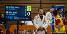 a man in a karate uniform stands in front of a scoreboard that says gustavo santos and bra gracie barra