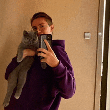 a man in a purple hoodie is holding a cat and taking a picture of himself