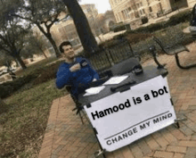 a man is sitting at a table with a sign that says hamood is a bot .