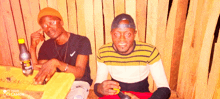 two men are sitting in front of a wooden wall with a bottle of sprite on the table