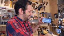 a man in a plaid shirt is holding a cell phone in front of a workbench with tools on the wall