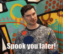 a man in a sweater says spook you later in front of a colorful wall