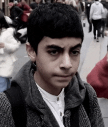 a young boy wearing a gray sweater and a white shirt looks at the camera