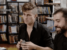 a man in a black shirt is holding a card in front of a bookshelf and the words love it are above him