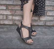 a woman wearing a black polka dot dress and black sandals stands in front of a brick wall