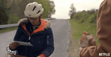 a woman wearing a helmet is reading a piece of paper next to a person holding a bag of bread