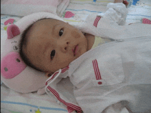 a baby is laying on a bed with a pink stuffed animal on his head and a white jacket with red stripes