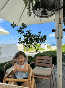 a little girl sits in a high chair under an umbrella on a beach