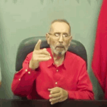 a man in a red shirt and glasses is sitting at a desk making a funny face .