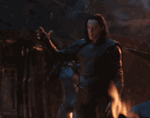 loki from avengers infinity war is holding a blue cube in his hand while standing next to a fire .