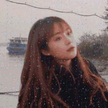 a woman with long brown hair is standing in front of a body of water with a boat in the background