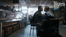 a man and a woman are sitting at a table in a store with the boys written on the ceiling