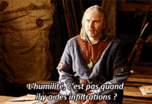 a man in a medieval costume is sitting on a wooden bench and says l' humilite