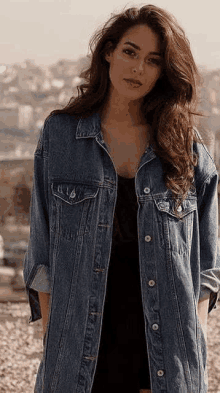 a woman wearing a long denim jacket and a black dress is standing in the dirt .