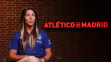 a woman in a blue shirt is sitting in front of a brick wall with the words atletico de madrid in red letters