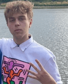 a young man is standing in front of a body of water wearing a white shirt with a drawing of a man on it .