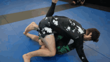 two men are wrestling on a blue mat and one of them is wearing a black and white shirt with white clouds on it