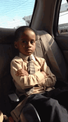 a young boy in a suit and tie sits in the back seat of a car with his arms crossed