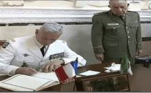 a man in a white suit is signing a document while another man stands behind him .