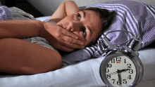 a woman covering her mouth while laying on a bed next to an alarm clock that shows the time as 7:15