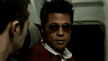 a man wearing sunglasses sits next to another man on an airplane