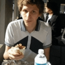 a young man is sitting at a table holding a sandwich and a bottle of water