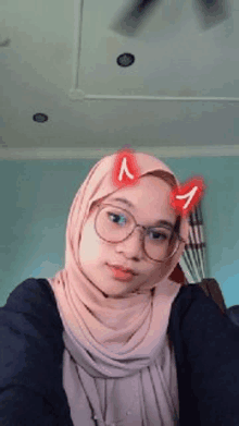 a woman wearing a hijab and glasses is taking a selfie with devil horns on her head .