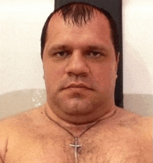 a shirtless man with a cross necklace around his neck looks at the camera