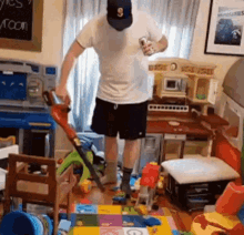 a man wearing a hat with the letter s on it is standing in a messy room holding a vacuum cleaner .