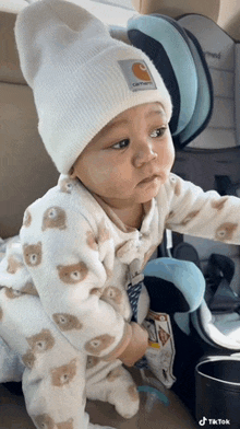 a baby wearing a white carhartt hat and a bear onesie is sitting in a car seat .