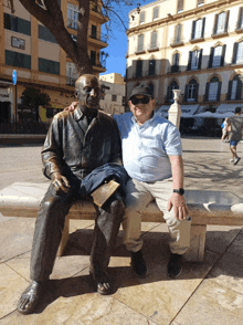 a man sits next to a statue of a man on a bench in front of a building that says " avenida "