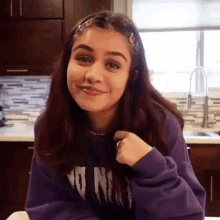 a woman in a purple sweatshirt is sitting at a table in a kitchen and smiling .