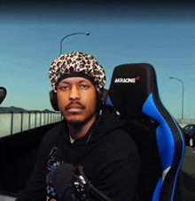 a man wearing a leopard print hat and headphones sits in a blue and black akracing chair