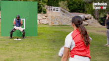 a woman in a baseball uniform is throwing a ball in front of a green screen that says jackass forever on it