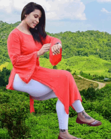 a woman in a red top and white pants is sitting on a hill looking at her watch