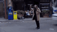 a woman in a tan coat is standing on a street in front of a yellow box that says recycling